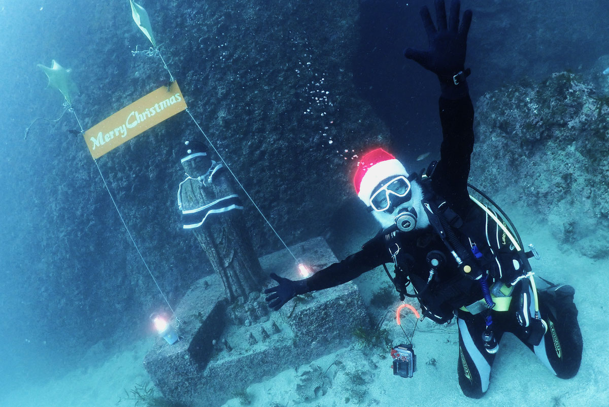水中のクリスマス飾りの前で手を振るサンタ姿のダイバー